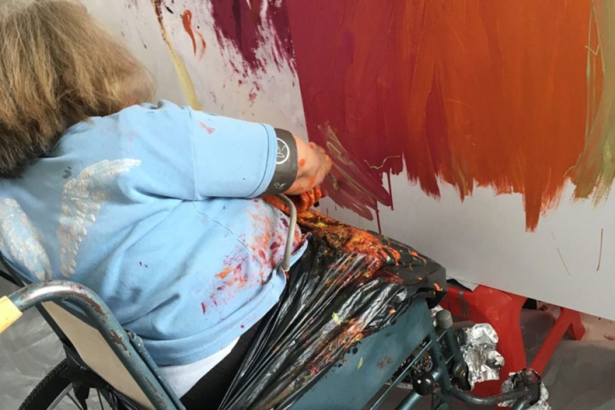Lady in Wheel Chair painting wall and all over her clothes with a sense of freedom although showing a form of disability. Art Therapy for Adults with Learning Disabilities