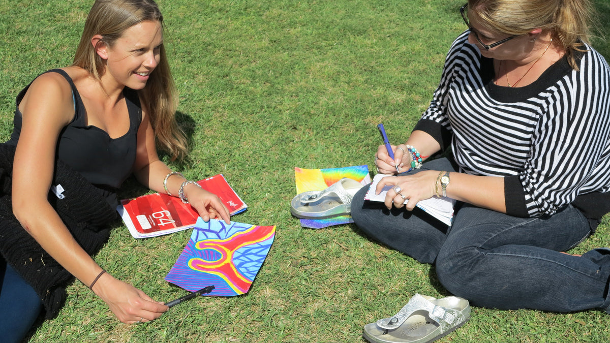 Two female students sitting on the grass with their drawings and taking notes.