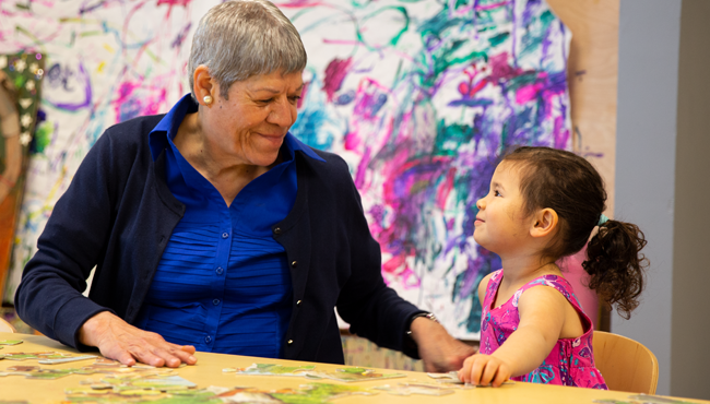 An older female art therapist and a young girl smile and connect in front of a large, vibrant, and messy wall painting created by children.