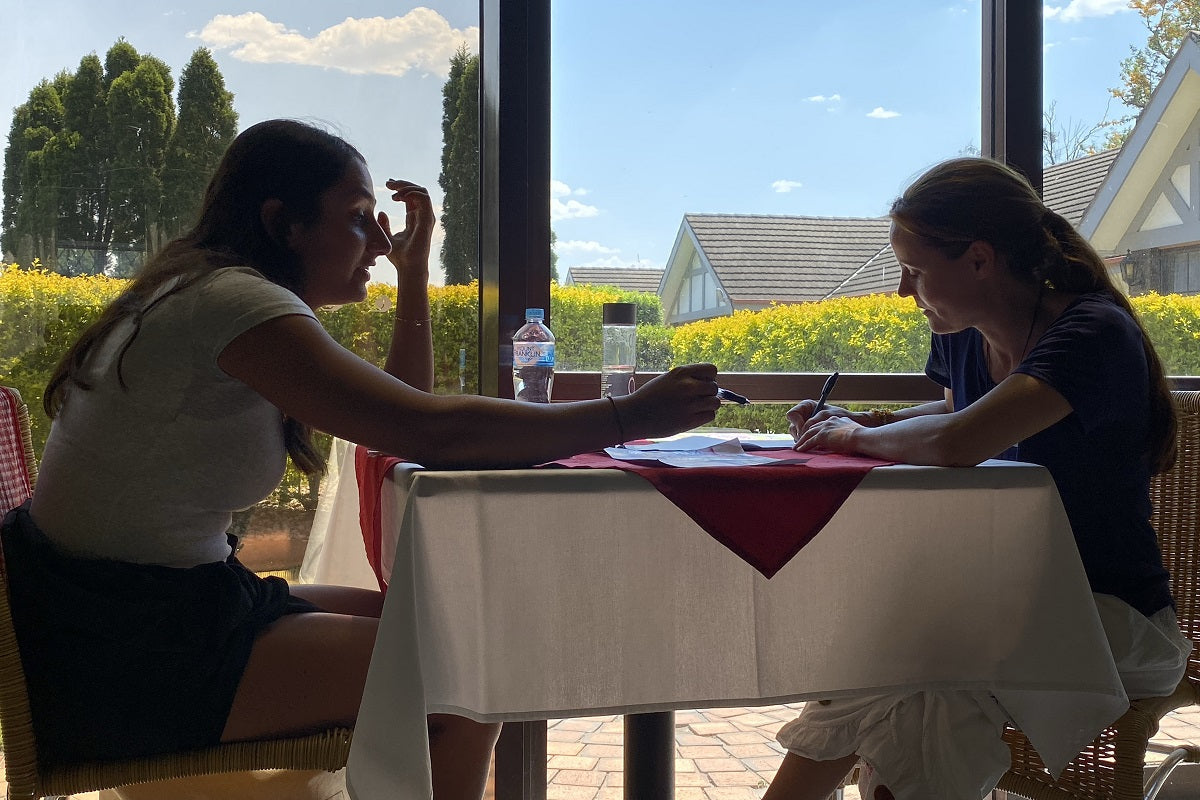 Two art therapy students talking at a table, one drawing while listening, with a calm outdoor view of trees and houses through the window.