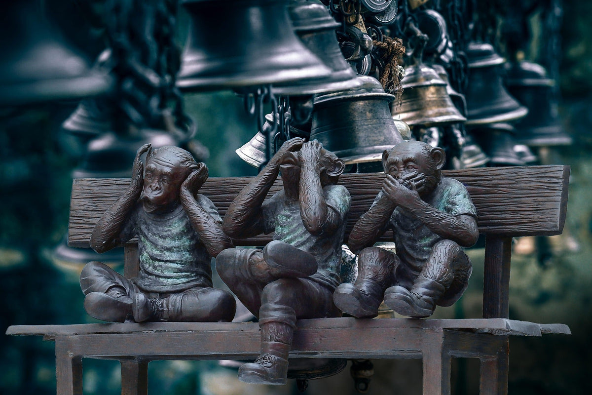 Three Wise Monkeys figurines sitting on a bench, depicting the "see no evil, hear no evil, speak no evil" proverb with bells in the background.