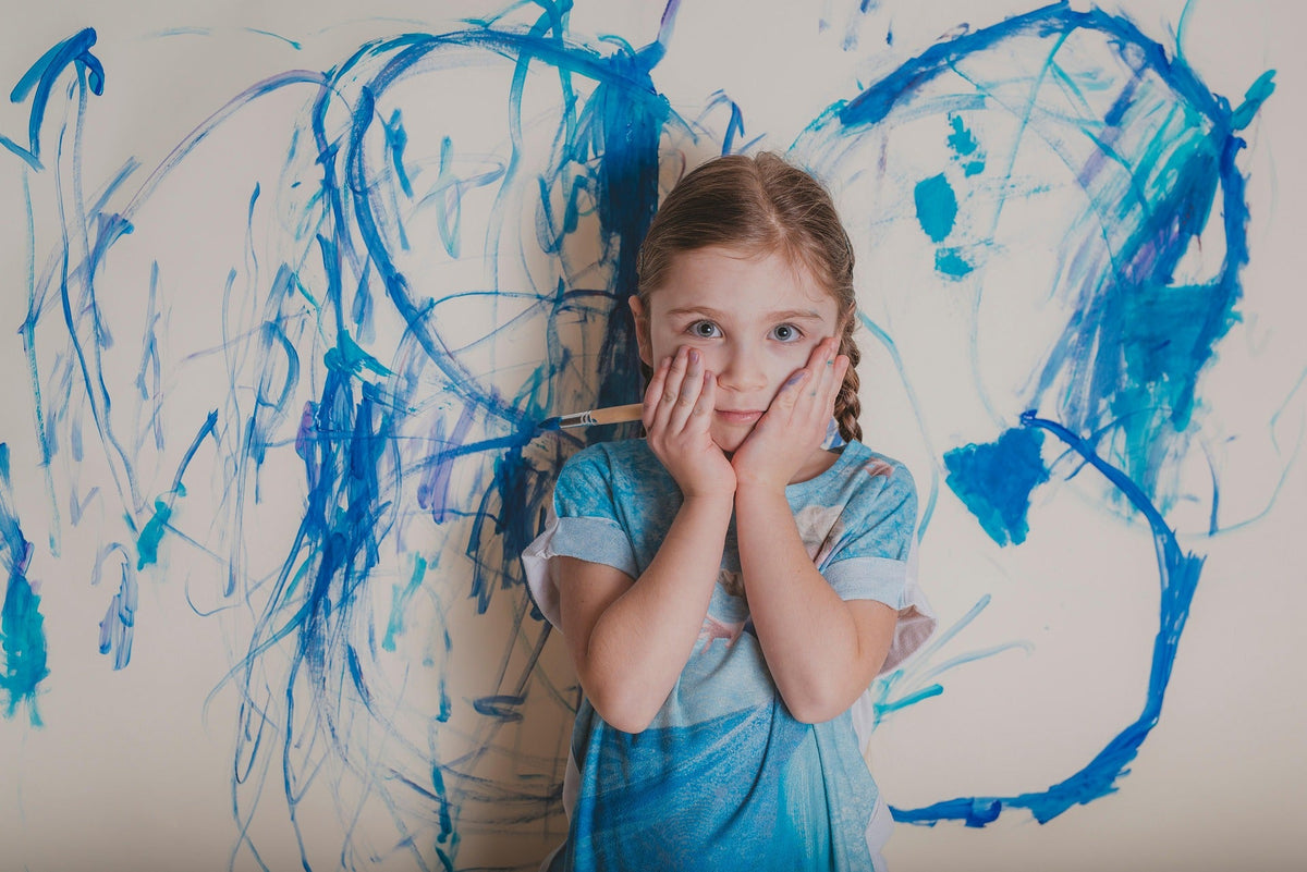 A young autistic girl holding a paintbrush stands in front of a wild, emotional blue abstract canvas she has started during an art therapy session