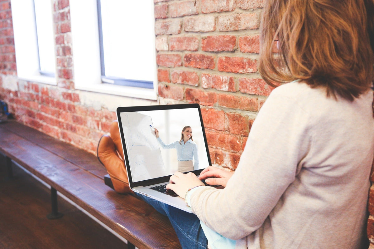 A woman sits on a bench in front of a brick wall while watching an online art therapy course on her laptop.